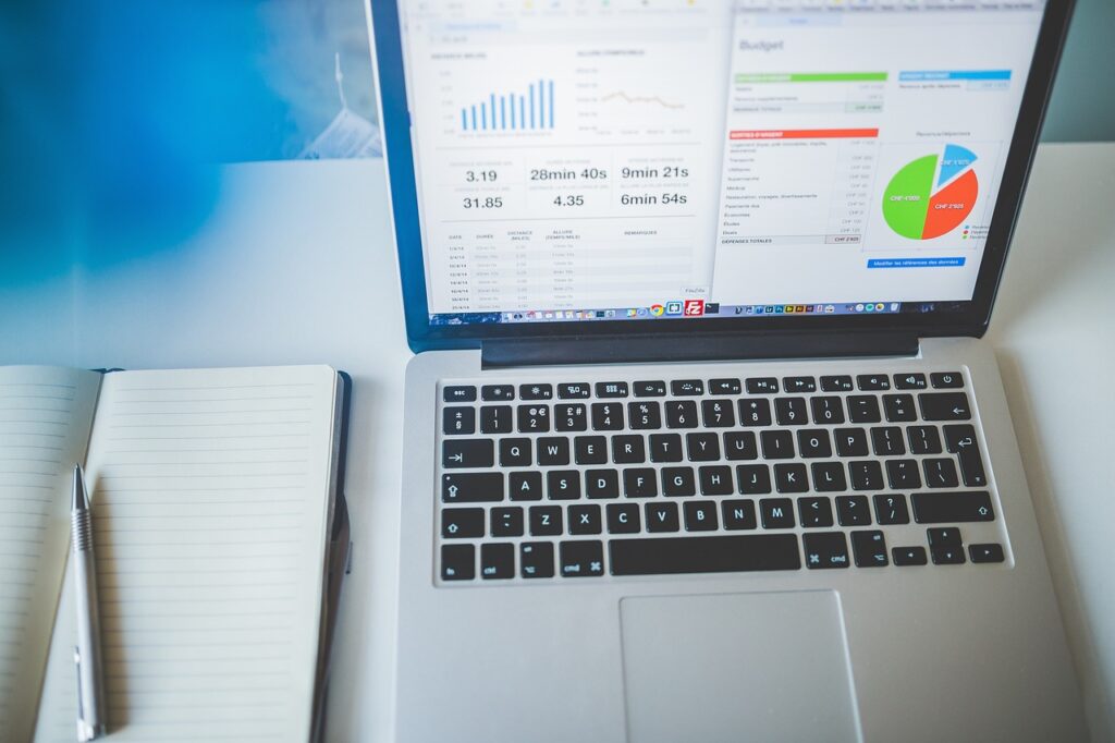 Laptop displaying business charts and budget data next to an open notebook and pen on a desk